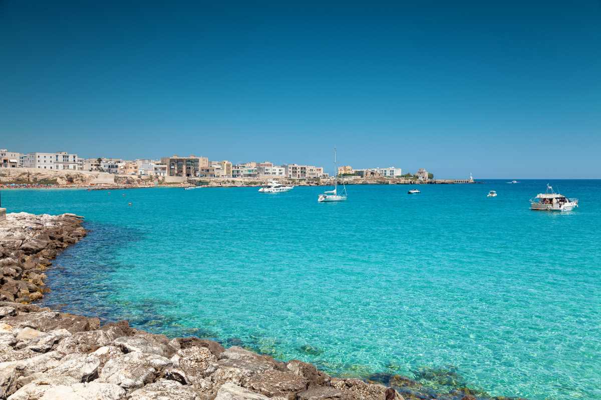 Foto panoramica del porto e del centro storico di Otranto in estate, con turisti e barche ormeggiate, cielo azzurro e colori vivaci